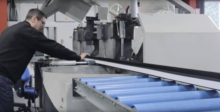 A person operates a large industrial machine with blue conveyor rollers, guiding a long black material—possibly used for sliding doors—through the equipment in a factory setting.