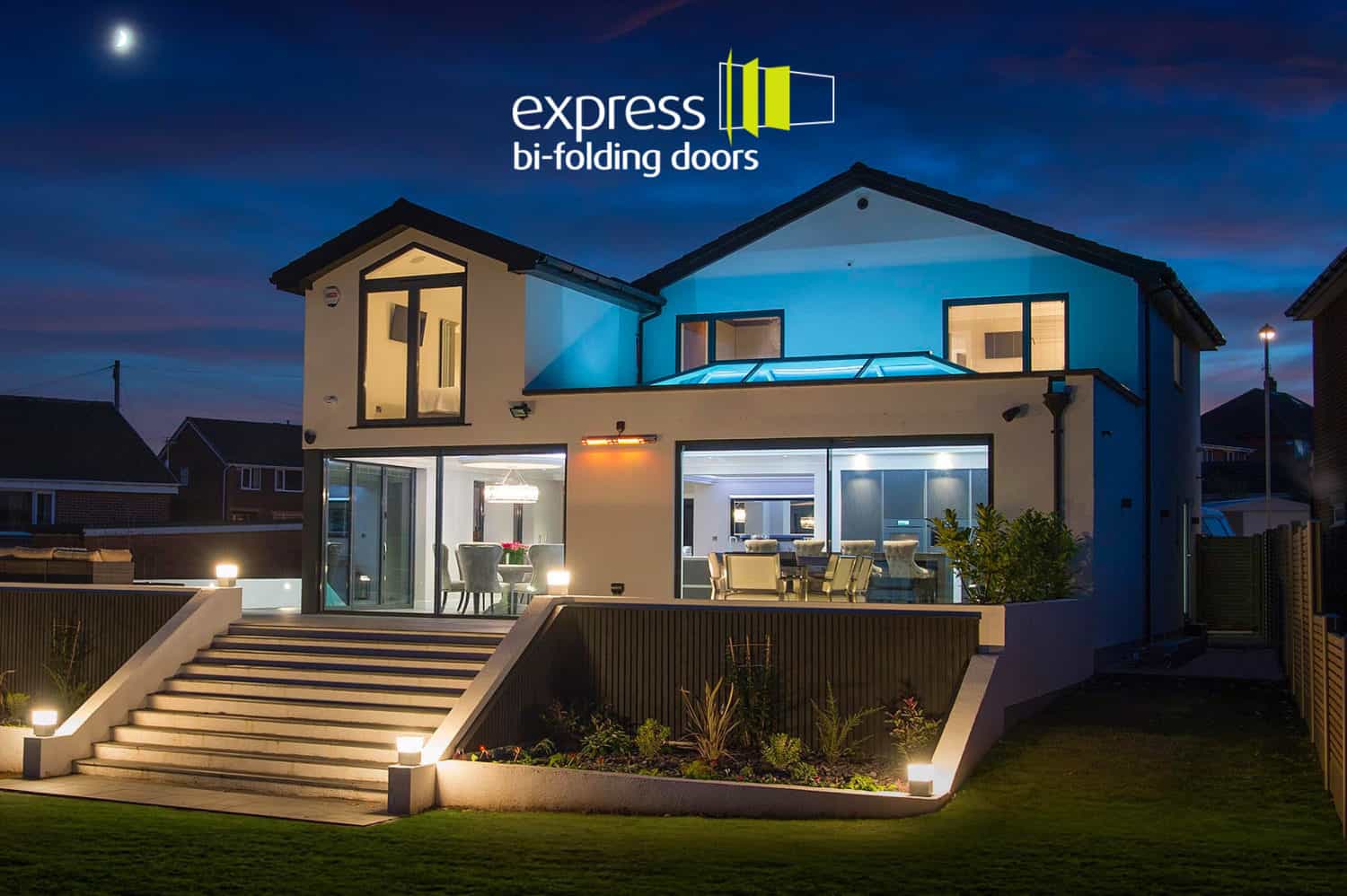 Modern two-story house at dusk with large glass bi-fold doors, sliding doors, outdoor lighting, landscaped garden, and the Express Bi-Folding Doors logo at the top. The interior is brightly illuminated.