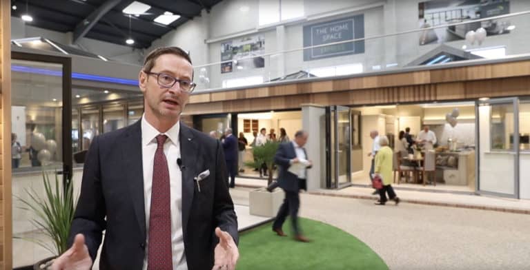A man in a suit and red tie speaks in a modern indoor showroom. People gather near a building display in the background, featuring bespoke glass solutions; a sign above reads The Home Space.