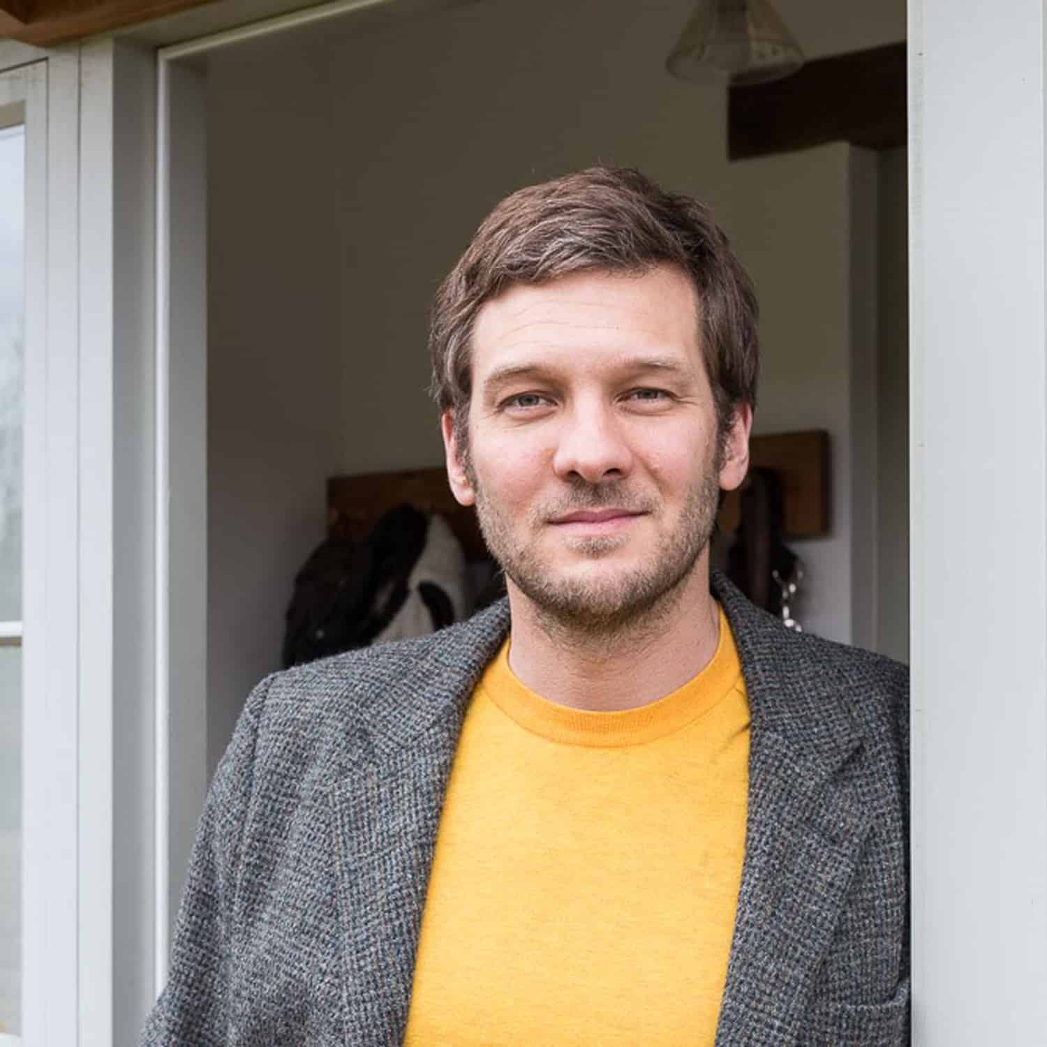 A man with short brown hair and light facial hair stands in a doorway with sliding doors, wearing a yellow shirt and a grey blazer, smiling softly at the camera.