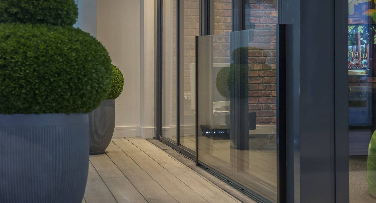 A modern sliding door is partially open, revealing large round green potted plants on a wooden deck. The scene includes reflections on the glass and a glimpse of a brick wall and colorful artwork inside.