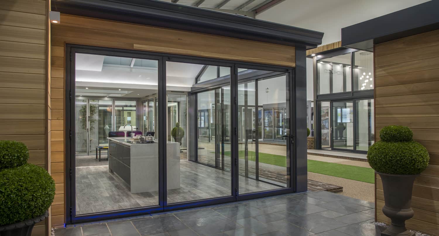 A modern showroom displays bespoke glass solutions, including large sliding doors, sleek kitchen cabinetry, and potted plants on a tiled floor, with natural wood paneling and overhead lighting creating a contemporary atmosphere.