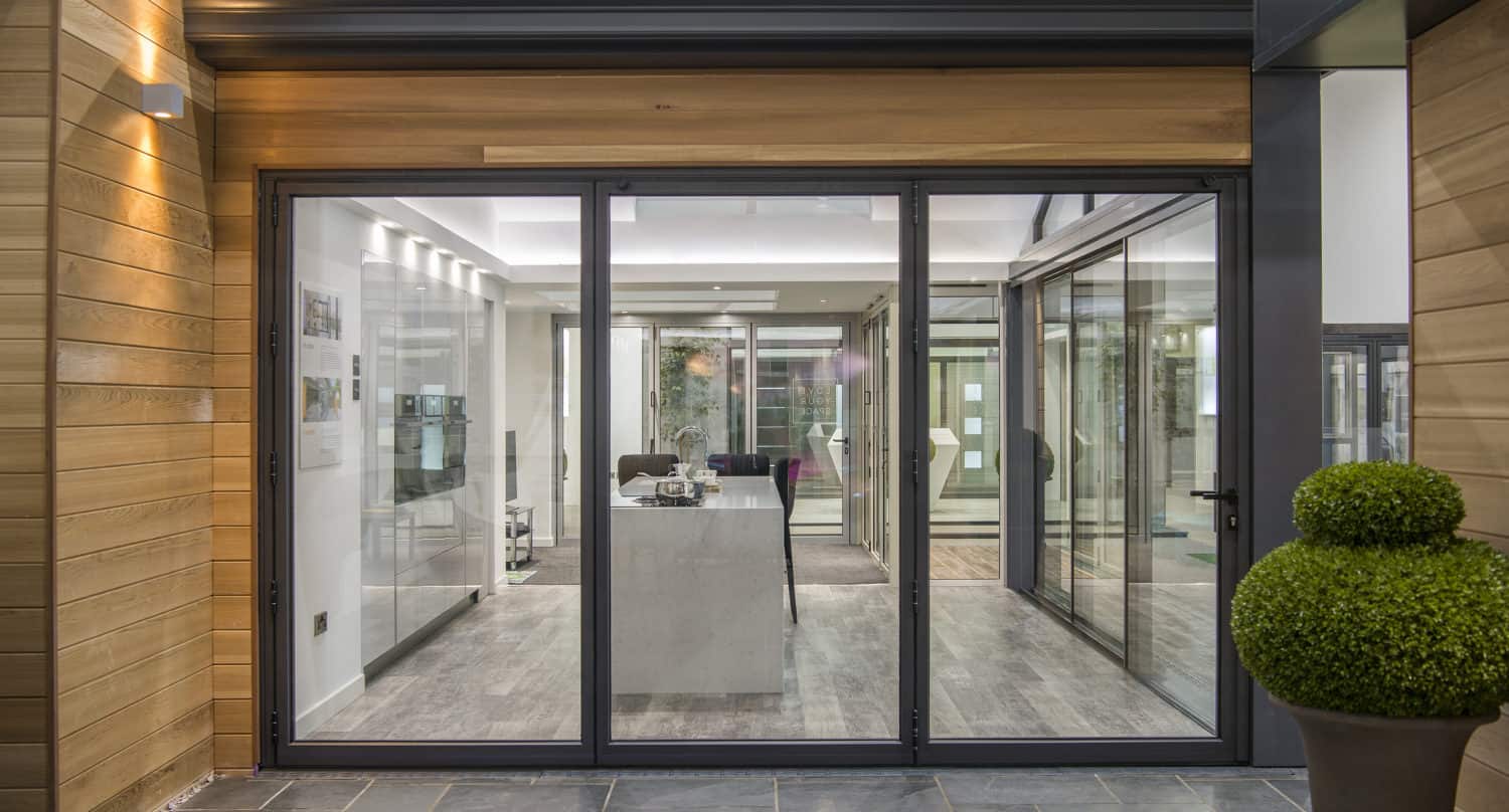 Modern sliding doors open into a bright, contemporary office space with wood and grey tile floors, a central desk, wall art, and a manicured round topiary plant placed outside, showcasing bespoke glass solutions for a stylish entryway.