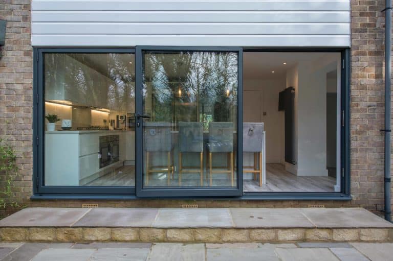 A modern home with large sliding glass doors, featuring bespoke glass solutions, opens to a kitchen and dining area. Four gray chairs sit at a wooden table; the kitchen has white cabinets and built-in appliances. Trees reflect in the glass.