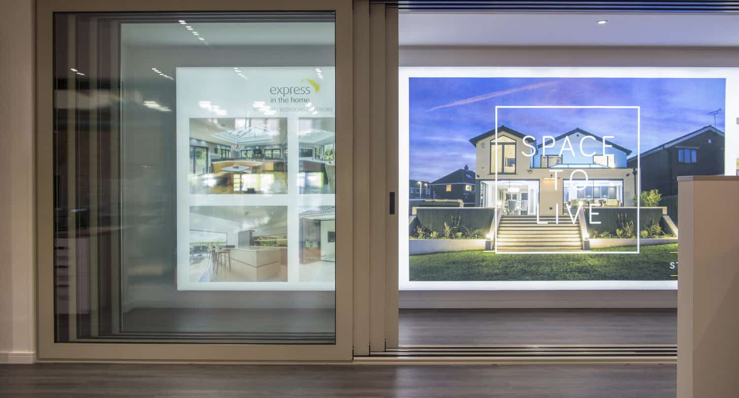 Modern interior with large bifold doors; behind them, illuminated displays feature house photos and the phrase “SPACE TO LIVE” over a picture of a contemporary home at dusk.