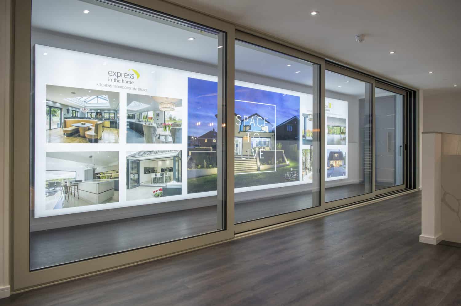 A modern showroom with large glass windows showcases illuminated posters of home interiors and exteriors, including bespoke glass solutions, on a white wall. The space features dark wood flooring and recessed ceiling lights.