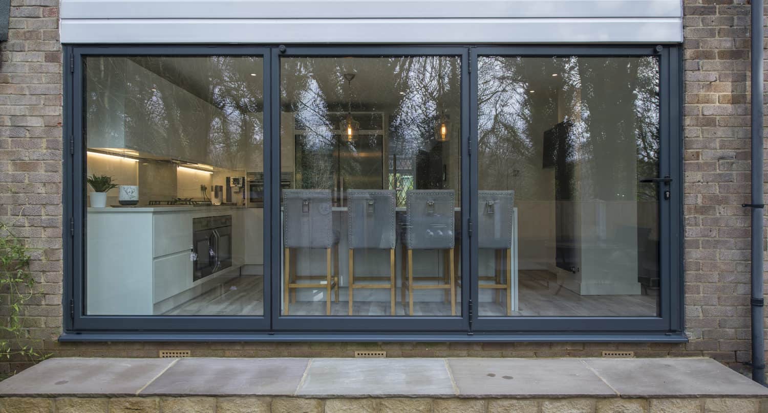 Large bifold doors reveal a modern kitchen and dining area with light grey chairs and wooden legs, white cabinets, and pendant lights. The view shows reflections of trees outside.