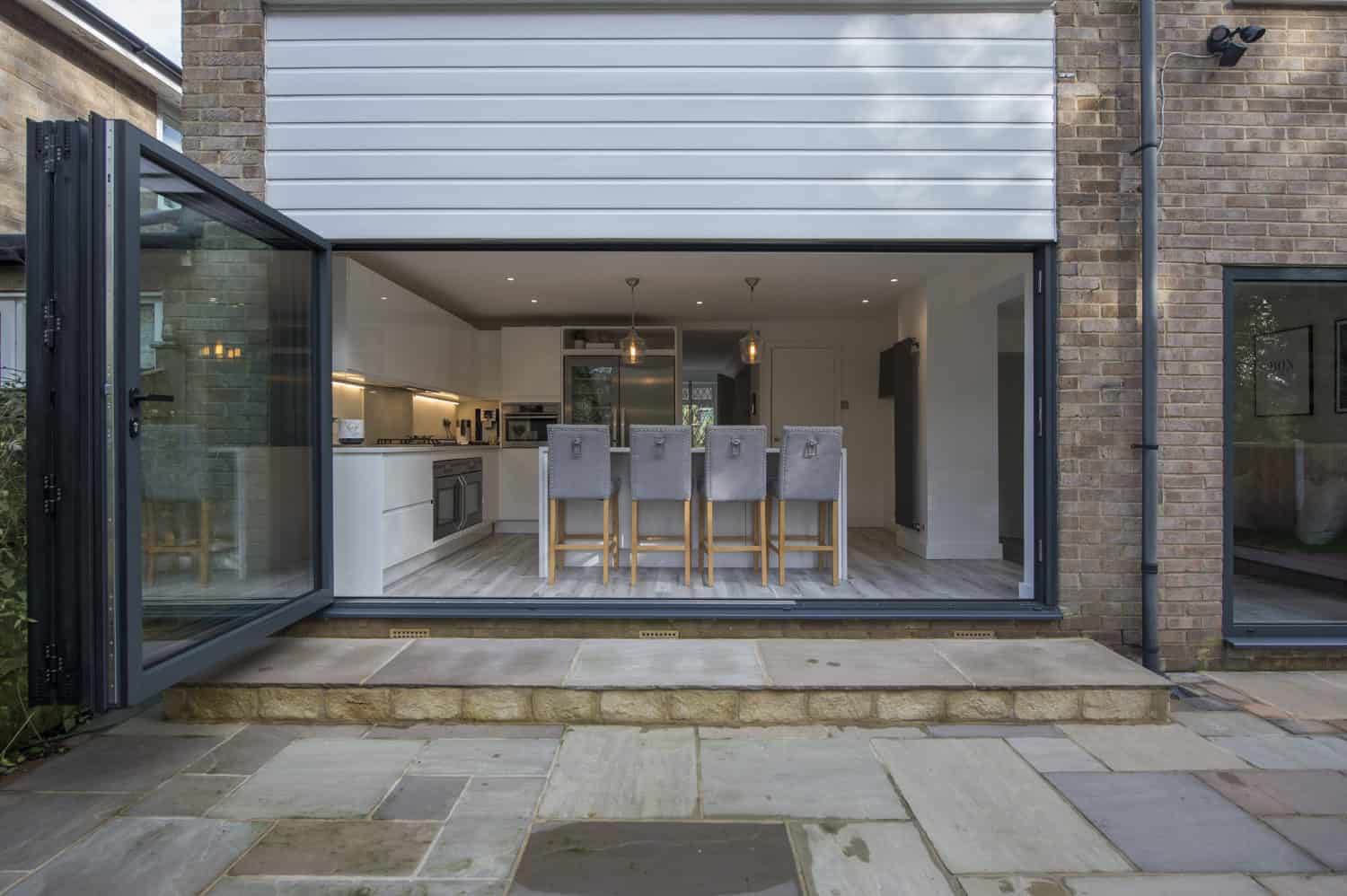 Modern open-plan kitchen and dining area with tall grey chairs, seen through wide open sliding doors from an outdoor stone patio. The space features contemporary decor, glass roofing, and plenty of natural light.
