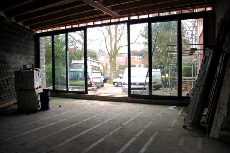 Interior view of a building under construction with large floor-to-ceiling windows and sliding doors; outside, vans and scaffolding are visible, along with trees and neighboring houses.