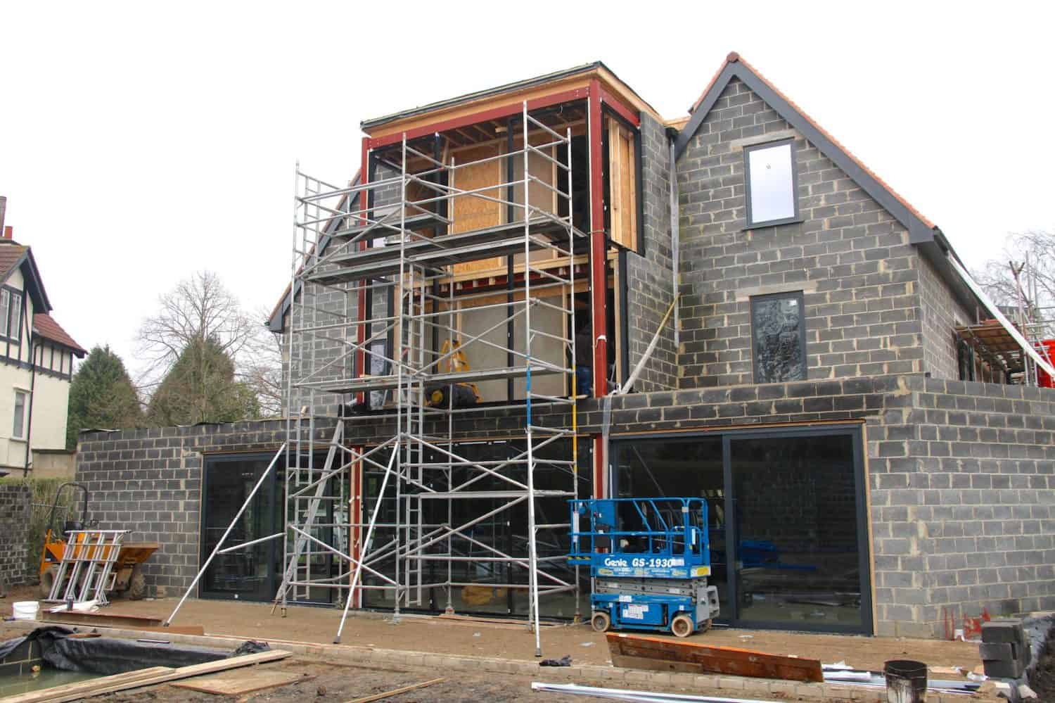 A modern house under construction with exposed brick walls, scaffolding, and a blue lift in front. The upper floor is being built, featuring bespoke glass solutions and sliding doors. Building materials are scattered around the site.