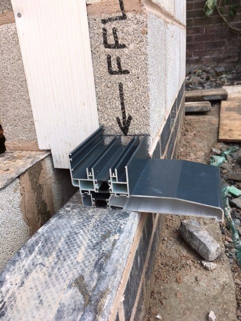 A close-up of a metal windowsill profile installed on a partially constructed brick and block wall, ideal for supporting sliding doors. An arrow and handwritten markings appear on the insulation panel above, with building materials and debris scattered below.
