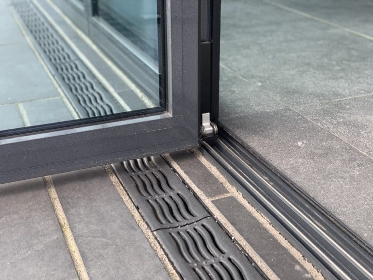 Close-up of a bespoke glass solution: a sliding glass door partially open, showing metal tracks embedded in a gray tiled floor with drainage grates nearby. The black door frame complements the modern, functional design.