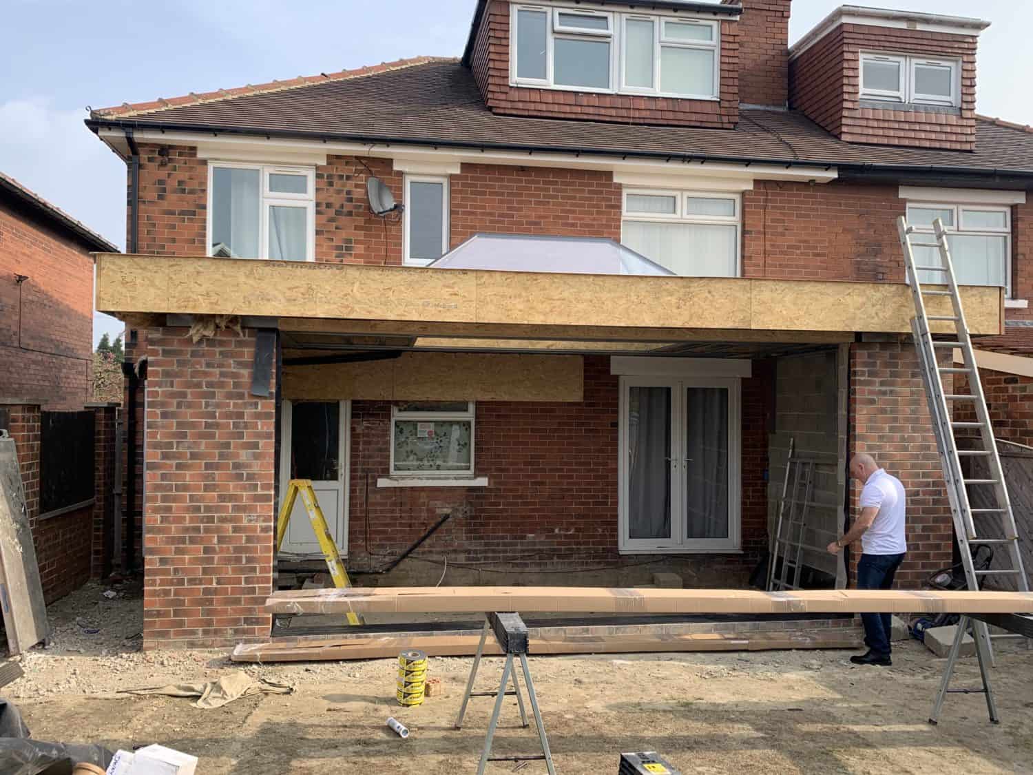 A man stands beside a partially built extension on a brick house. Wooden beams and construction materials are scattered around, with ladders leaning against the house. The house’s sliding doors and windows are visible at the back.