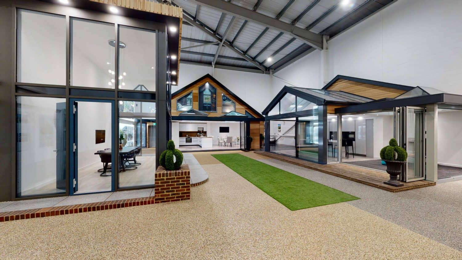 Modern indoor showroom displaying model house exteriors with large glass windows, sliding doors, and skylights, featuring artificial grass, potted plants, and a high ceiling with exposed beams.