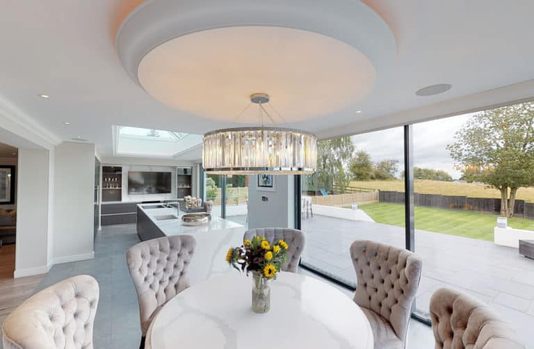 Modern dining room with a round table and tufted chairs, a vase of sunflowers, and a chandelier overhead. Large windows with bespoke glass solutions reveal a spacious patio and green backyard with trees.