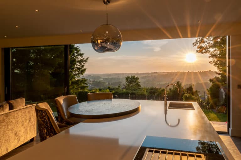Modern kitchen with a large island, sink, and pendant light, opening to a patio through bifold doors with bespoke glass solutions. Sunlight streams in at sunset, filling the space with a warm glow and framing views of trees and hills.