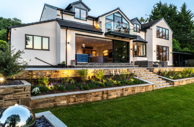 Modern two-story house with large windows and bespoke glass solutions, illuminated at dusk. The home features a landscaped garden, stone retaining walls, outdoor seating, and neatly trimmed grass in the foreground.