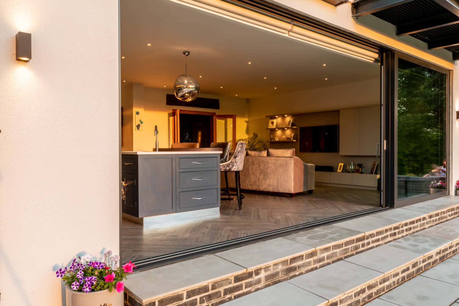 Open sliding bifold doors reveal a modern kitchen and living room with contemporary furniture, a kitchen island, and a hanging light fixture. Potted flowers sit outside on the stone patio.