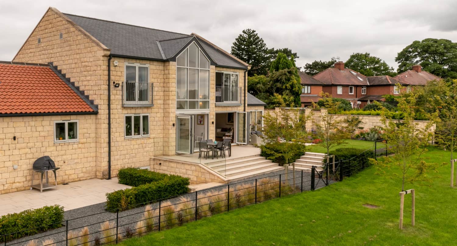 A modern stone house with large glass windows and bifold doors, an outdoor patio with seating, a barbecue grill, and a neatly landscaped lawn, surrounded by hedges and a black metal fence on a cloudy day.