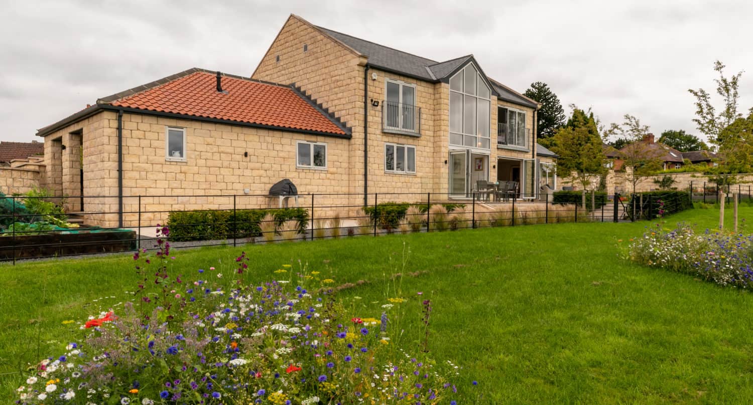 A modern stone house with bespoke glass solutions, including large windows and sleek bifold doors, sits on a spacious green lawn with colorful wildflowers in the foreground, under a cloudy sky.