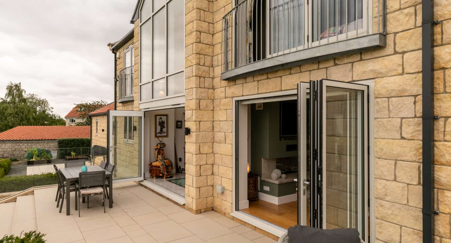 Modern stone house with large bifold doors opening to a spacious patio, featuring outdoor dining furniture and views of greenery and red-roofed buildings in the background under a cloudy sky.