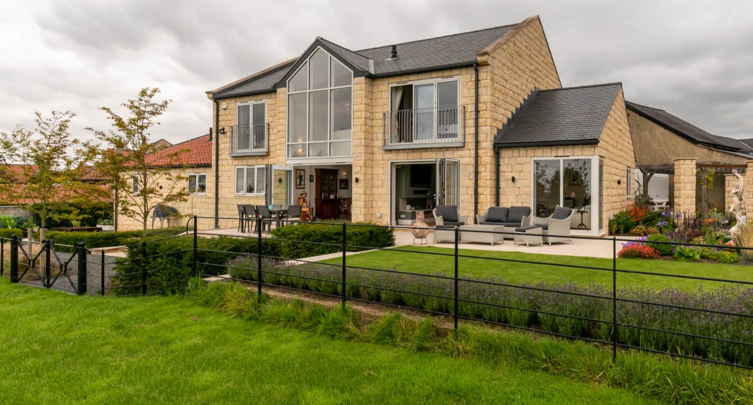 Modern two-story stone house with large windows and sliding doors, a spacious patio with outdoor seating, and well-maintained green lawn, surrounded by a black metal fence and landscaped garden beds under a cloudy sky.