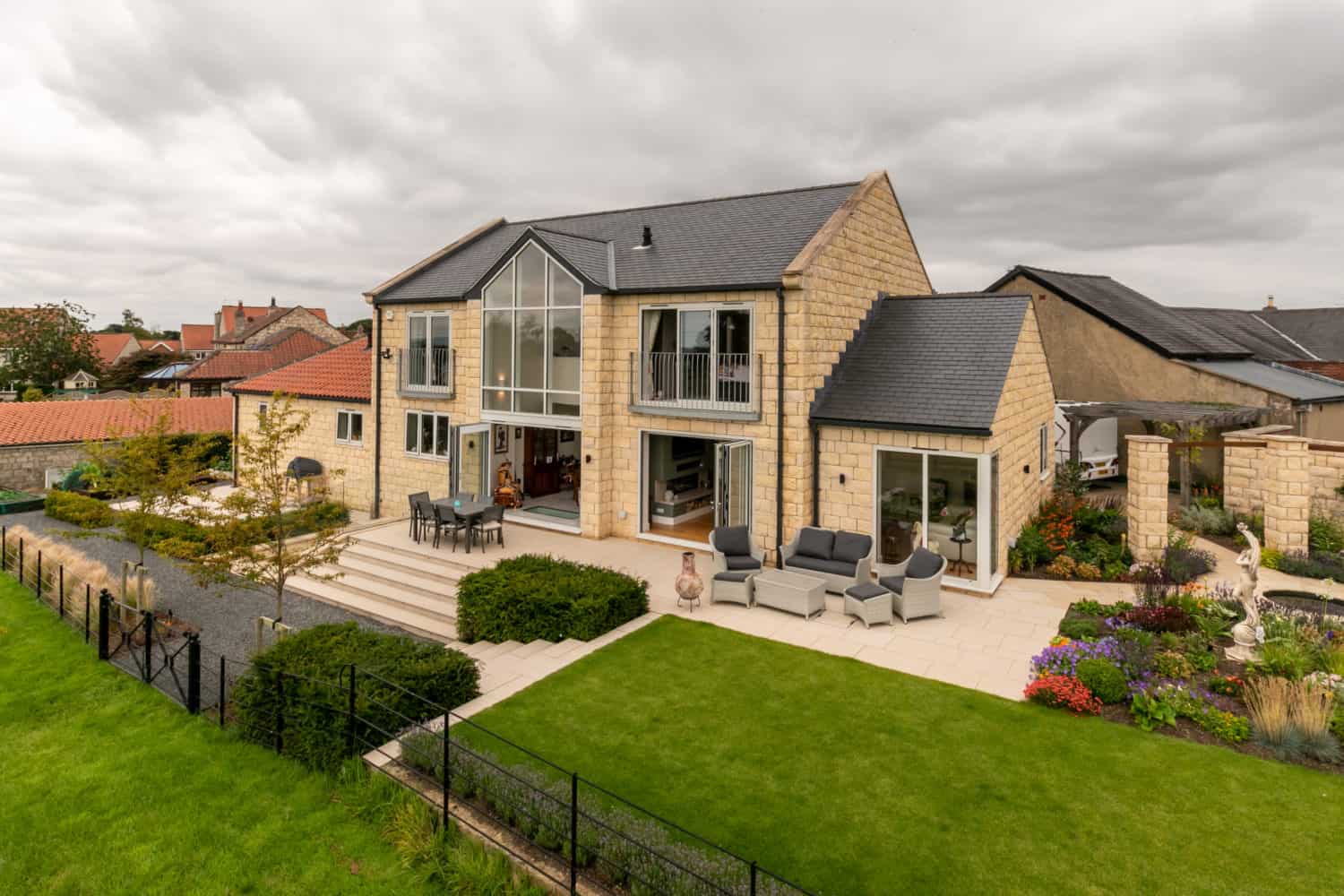 Modern stone house with large windows, bifold doors, a balcony, and a spacious patio featuring outdoor seating. The neatly landscaped garden includes lawns, flower beds, and a gravel path under a cloudy sky.