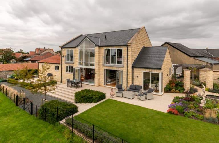 Modern stone house with large windows, bifold doors, a balcony, and a spacious patio featuring outdoor seating. The neatly landscaped garden includes lawns, flower beds, and a gravel path under a cloudy sky.