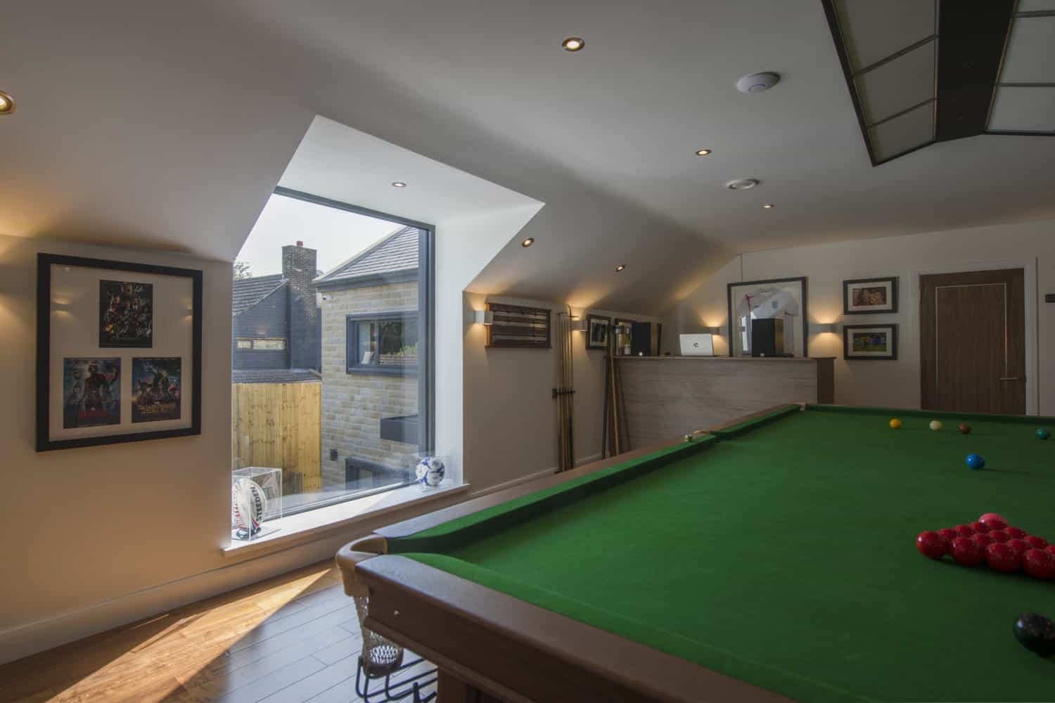 A well-lit attic game room with a green snooker table, framed pictures on white walls, wooden floors, and a sloped ceiling featuring spotlights and bespoke glass solutions that let in natural light from a large window overlooking neighboring buildings.