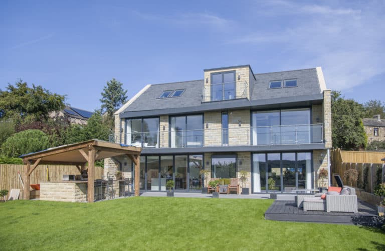 Modern three-story house with large glass windows and balconies, stone and wood exterior, stylish sliding doors, outdoor patio, covered seating area, and a well-kept lawn under a blue sky.