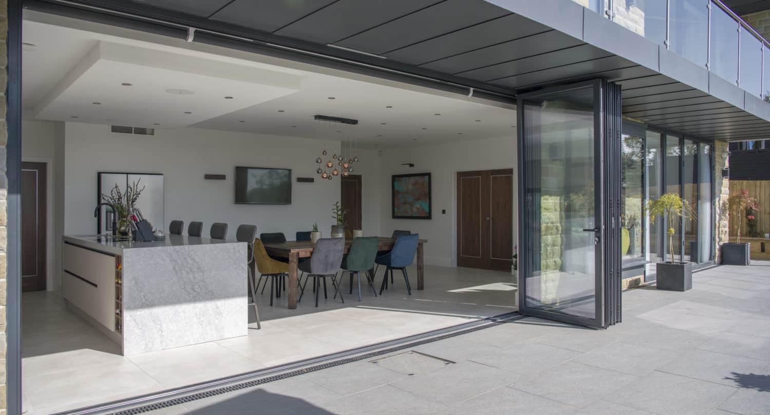 Modern open-plan kitchen and dining area with bespoke glass solutions, including large glass folding doors opening to a patio, marble island, colorful chairs, and sleek furnishings in a bright, spacious setting.