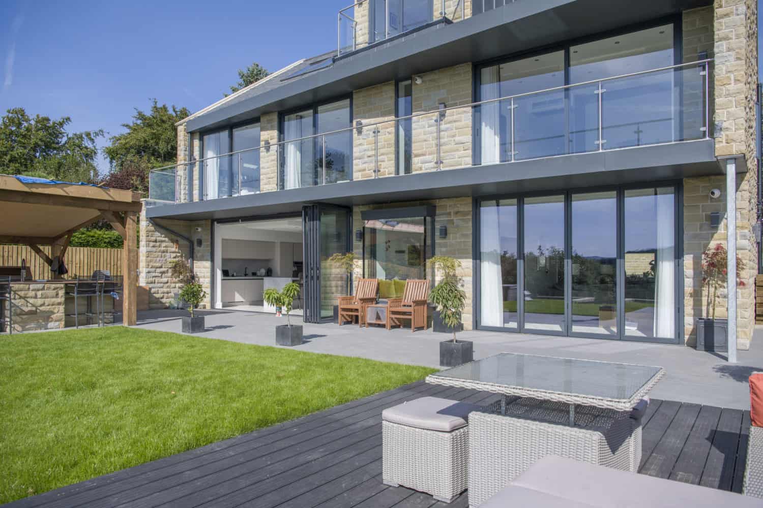 Modern house with large glass windows, bespoke glass solutions, balconies, stone exterior, and outdoor patio areas with seating and dining furniture, overlooking a well-kept lawn and a deck under a clear blue sky.