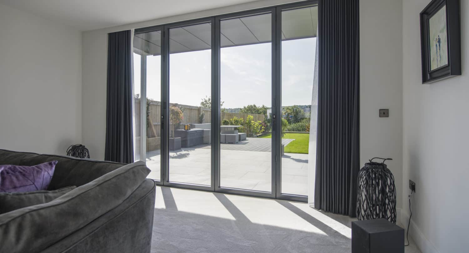 A modern living room with large floor-to-ceiling sliding doors that open to a patio with outdoor seating and a green lawn. Dark curtains frame the glass doors, and a gray sofa is partially visible inside.