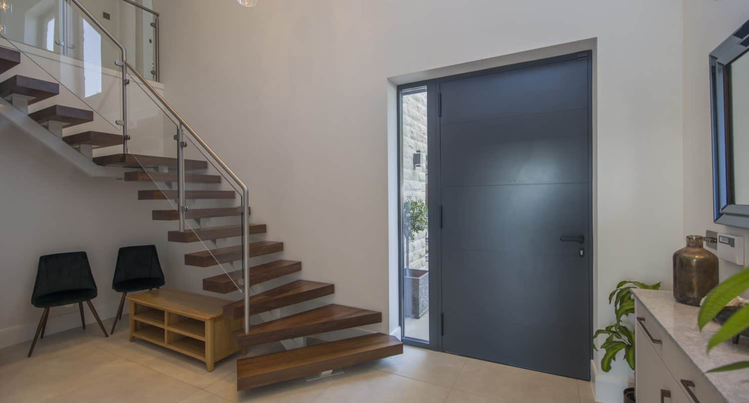 Modern entryway with floating wooden stairs, a large dark door, glass railing, two black chairs, and a wooden bench. Light fills the space from high windows and bespoke glass solutions, creating a clean, open atmosphere.