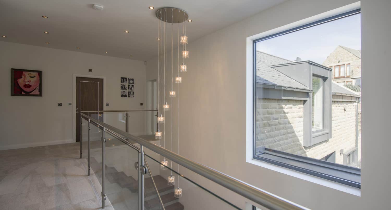 Modern hallway with glass railing, hanging pendant lights, large window with view of stone buildings, and wall art including a portrait and family photos. Neutral walls and floors plus bifold doors create a bright, airy atmosphere.