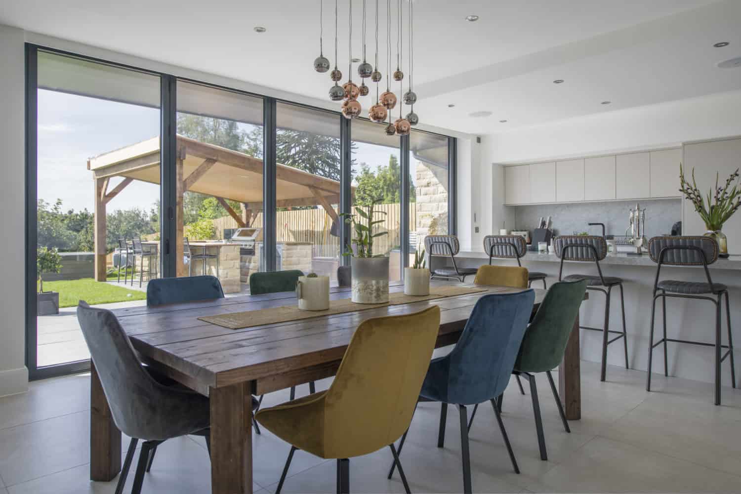 Modern dining and kitchen area with a large wooden table, colorful upholstered chairs, pendant lights, and bespoke glass solutions framing the view of a garden patio through floor-to-ceiling glass doors.