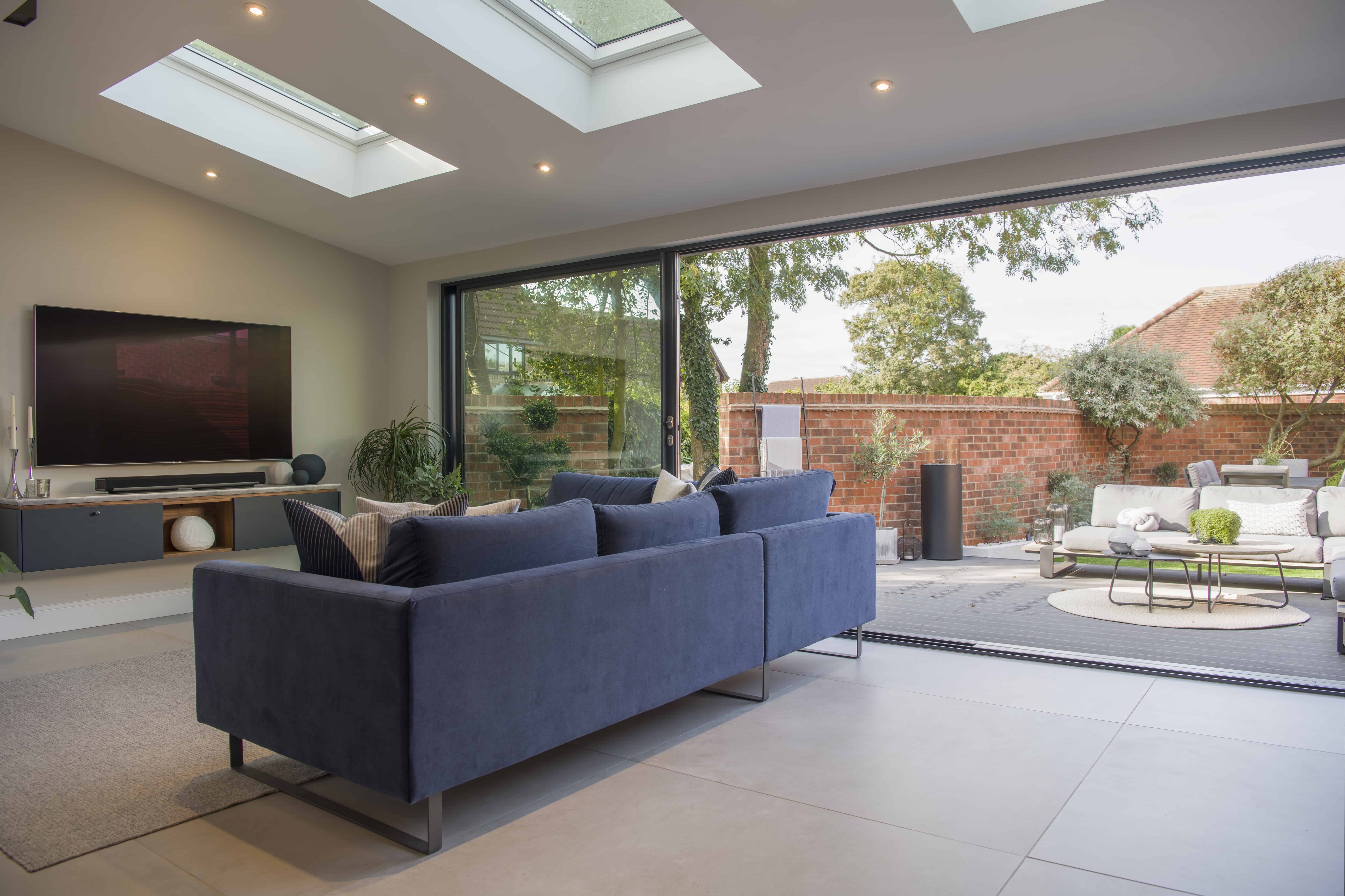 Modern living room with a blue sofa facing a wall-mounted TV, large sliding doors open to a patio with outdoor seating, skylights above providing natural light, and lush greenery outside.