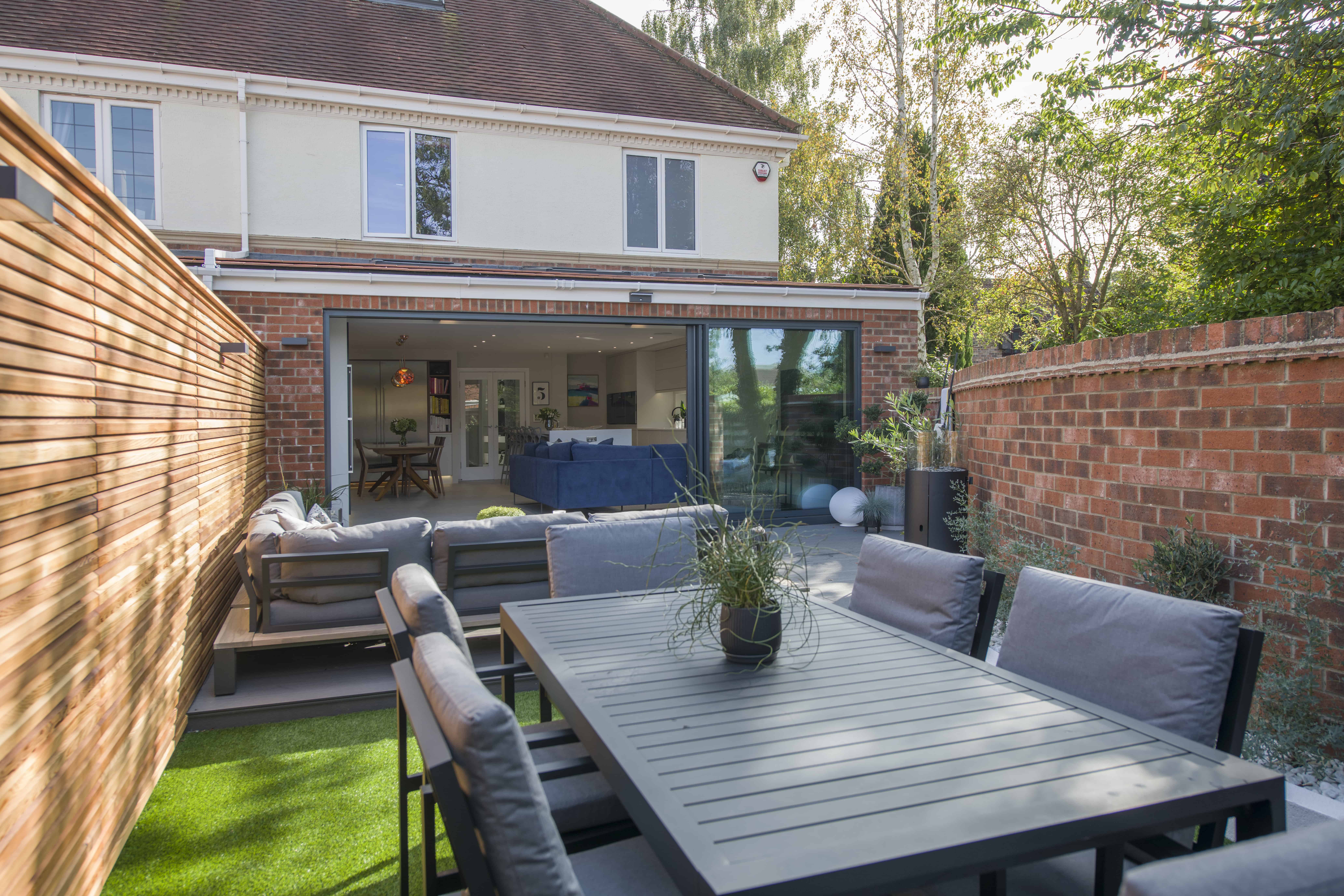 Modern backyard with a patio dining set, outdoor sofas, potted plants, artificial grass, and a brick fence. Bespoke glass solutions feature large sliding glass doors opening to the living area of the spacious house.