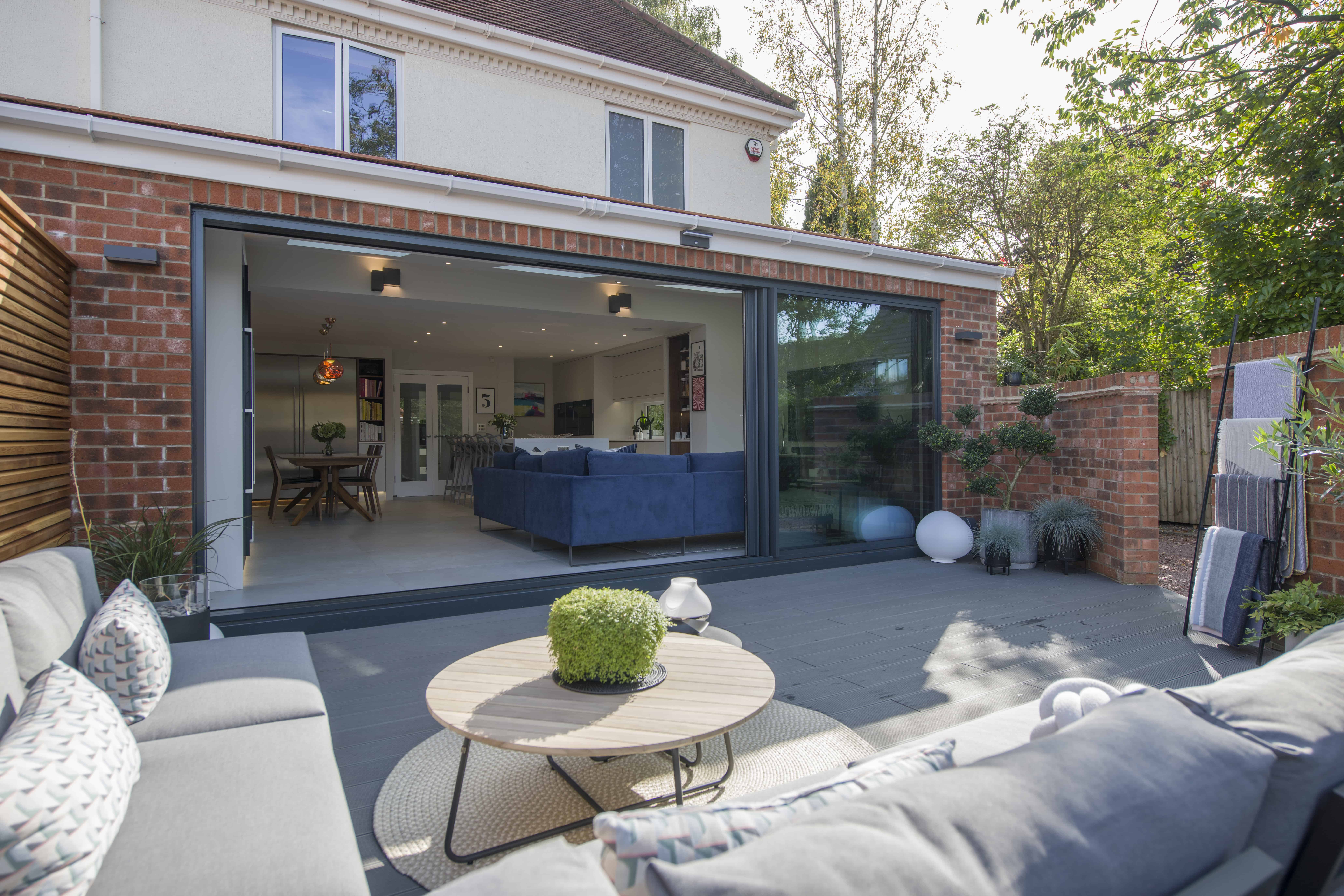 Modern patio with gray outdoor sofas and round coffee table overlooks bifold doors leading to a spacious living area with a blue sectional sofa inside a contemporary brick and white house. Lush greenery surrounds the space.