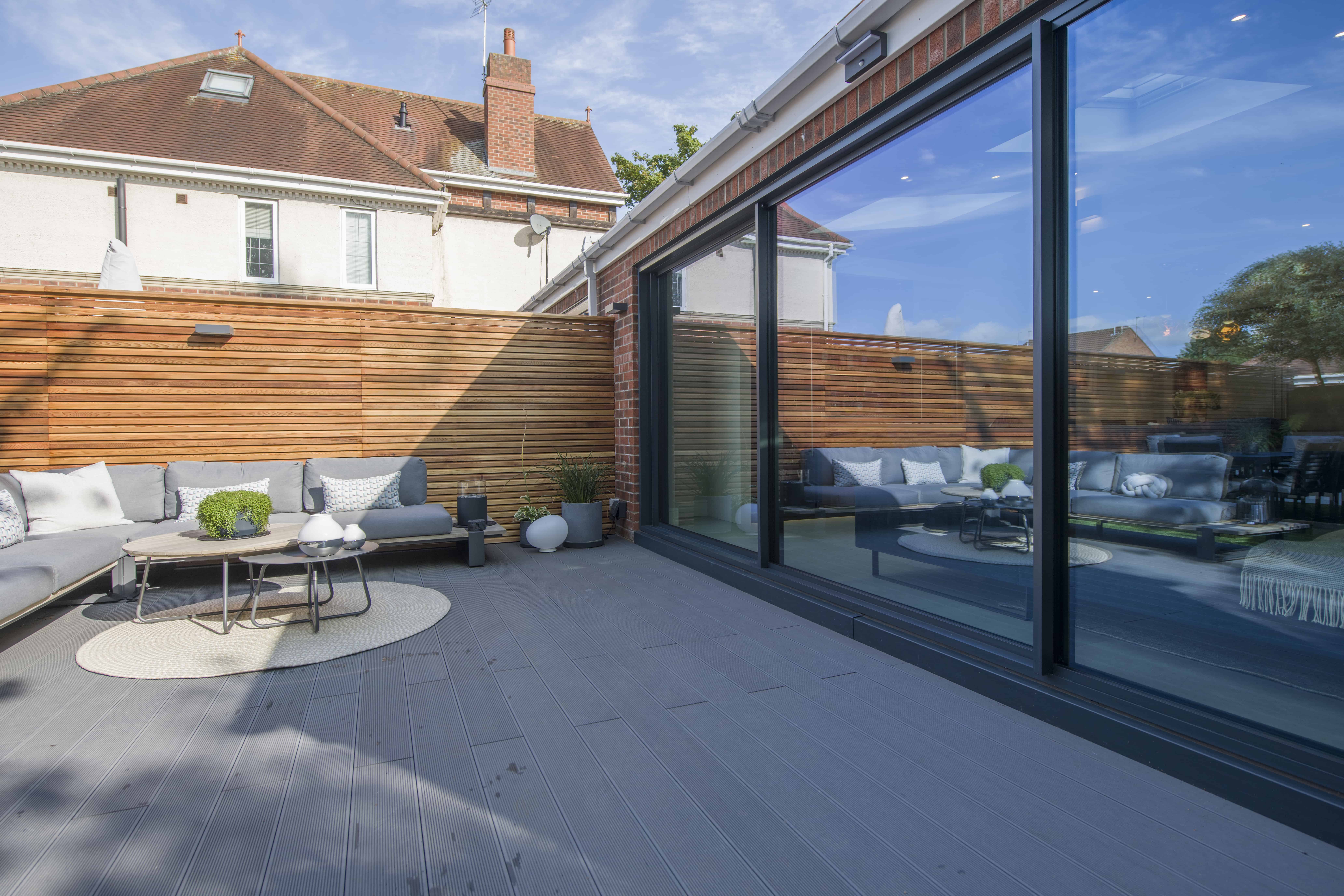Modern patio with gray decking, outdoor sofas, and tables. Wooden privacy fence surrounds the space. Large sliding doors reflect the stylish seating area and greenery. Sunny weather with blue sky above.