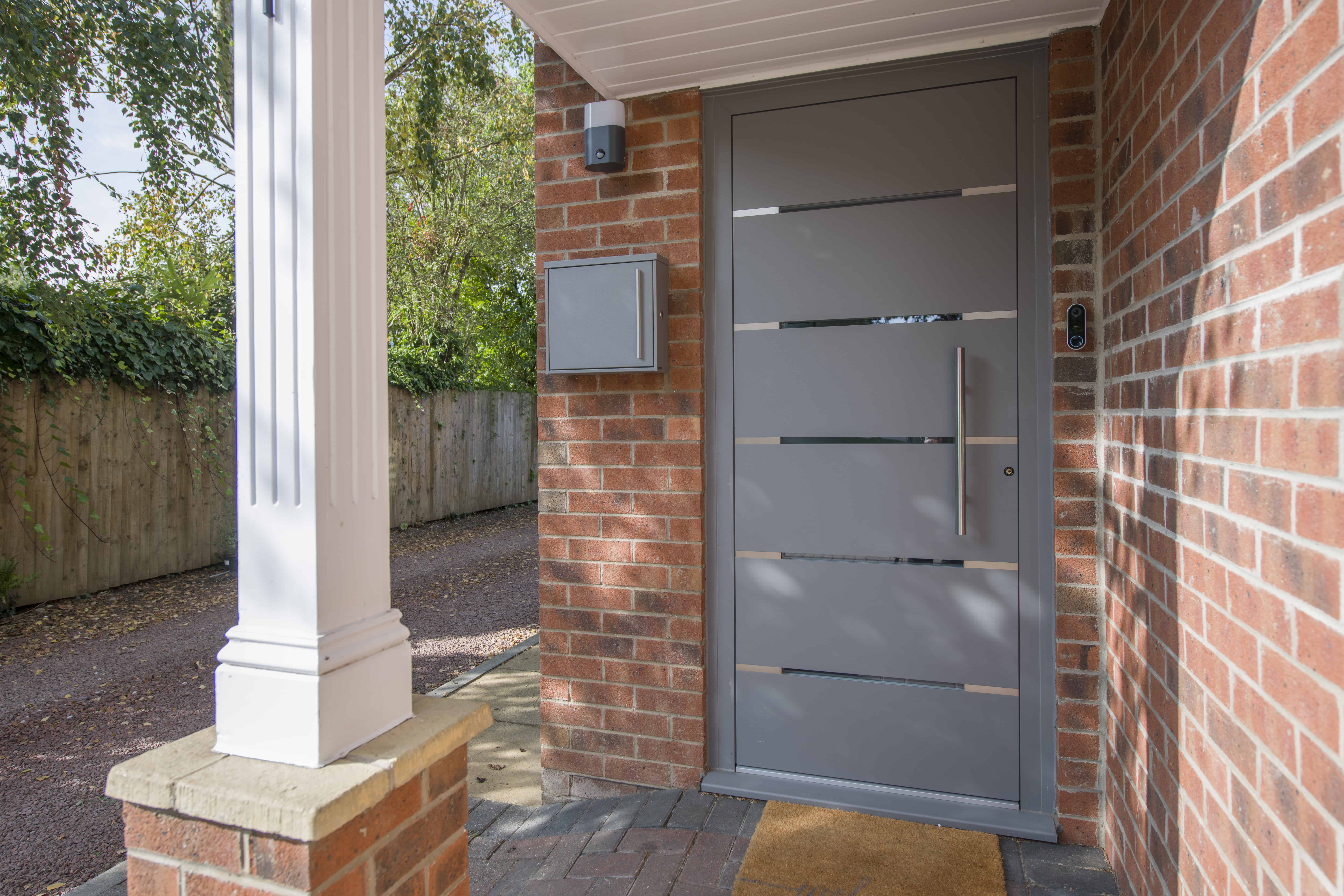 Modern gray front door with horizontal glass panels, a showcase of bespoke glass solutions, set in a brick house exterior; a mailbox is mounted on the wall, a column stands nearby, and a welcome mat is on the paved entryway.
