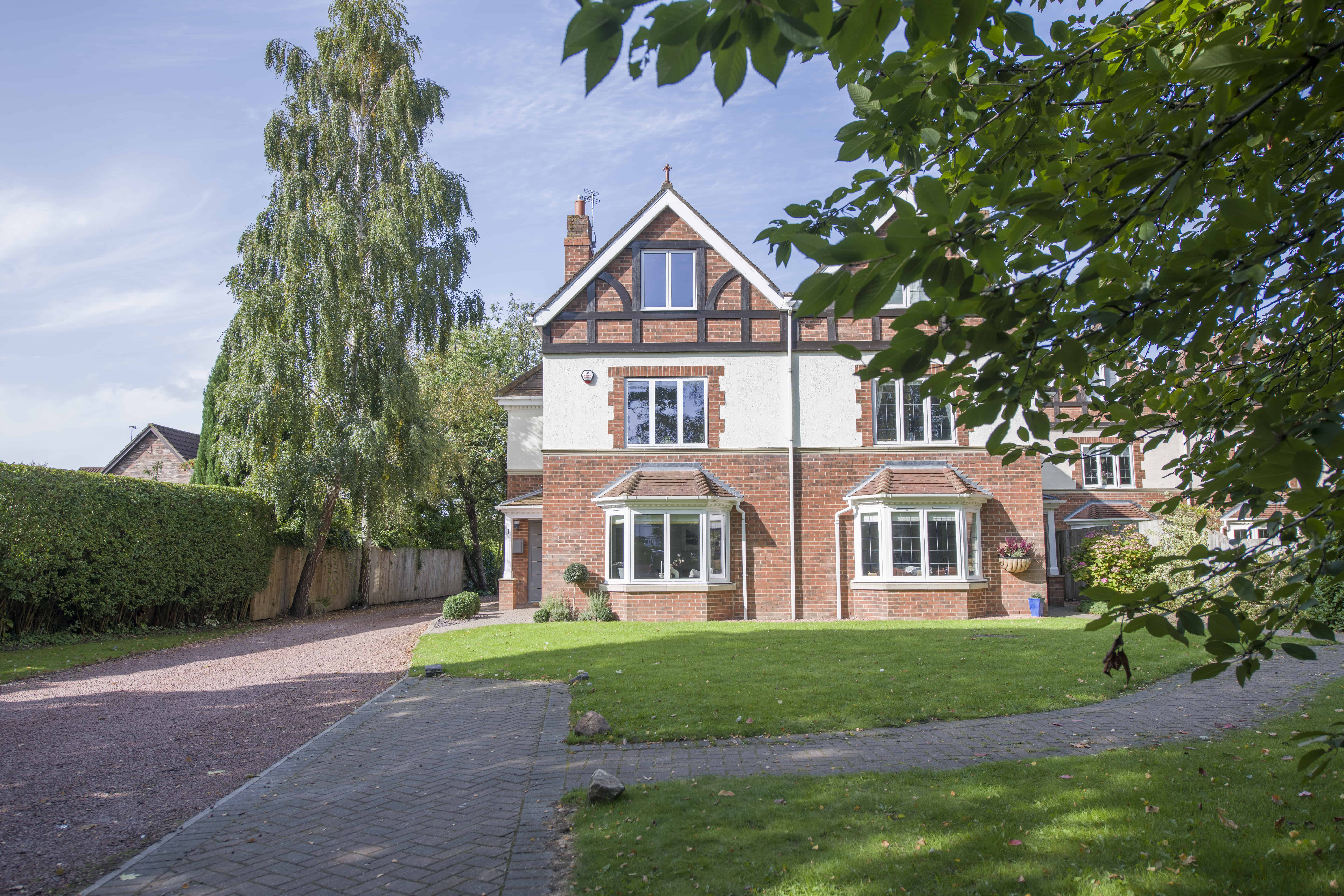 A large, two-story brick and white half-timbered house with bay windows and sliding doors, set among trees and greenery, with a paved driveway curving past a well-kept lawn under a sunny sky.