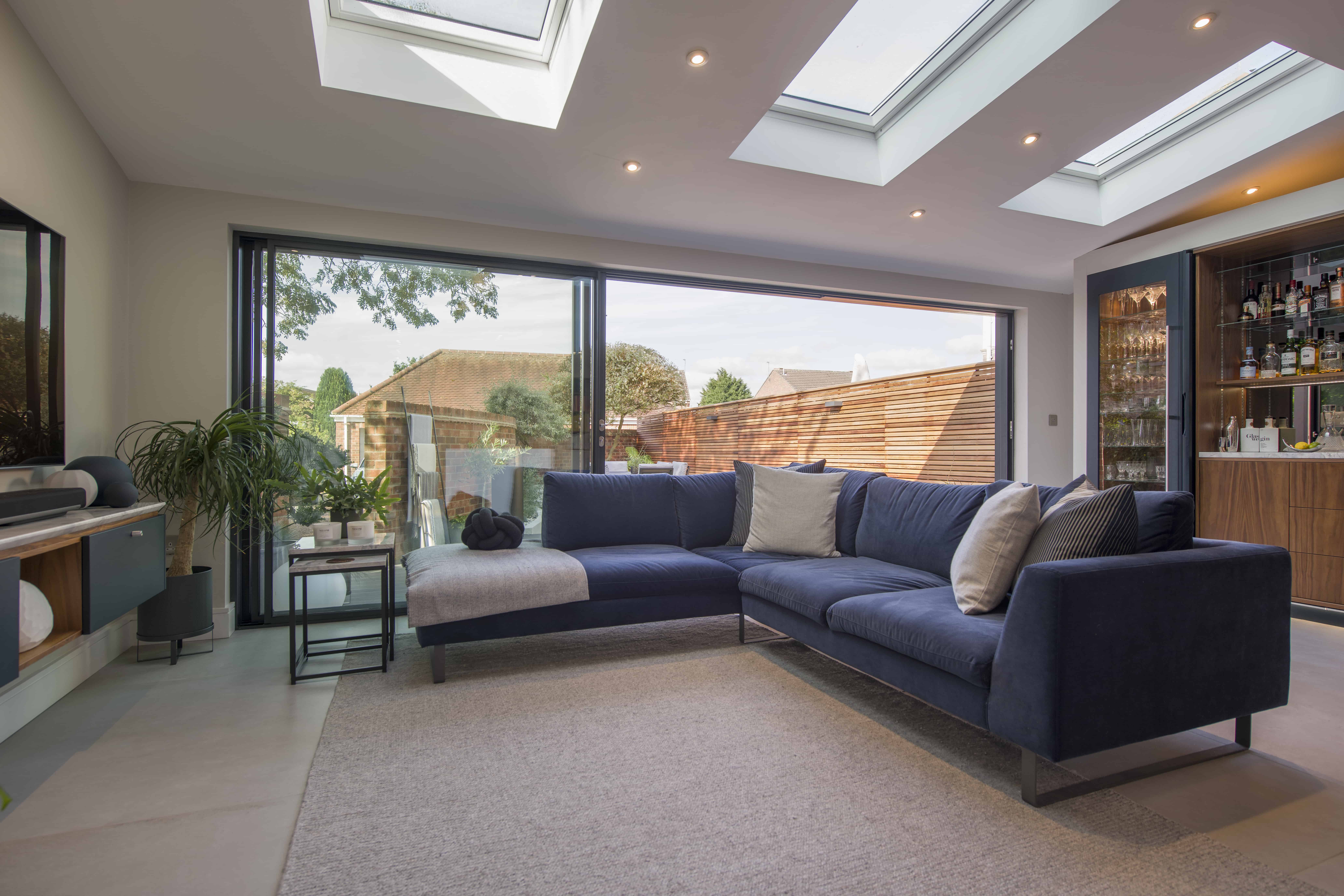 Modern living room with a large blue sectional sofa, neutral rug, indoor plants, and expansive bifold doors opening to a patio. Skylights and recessed lighting brighten the space. A home bar is visible on the right.