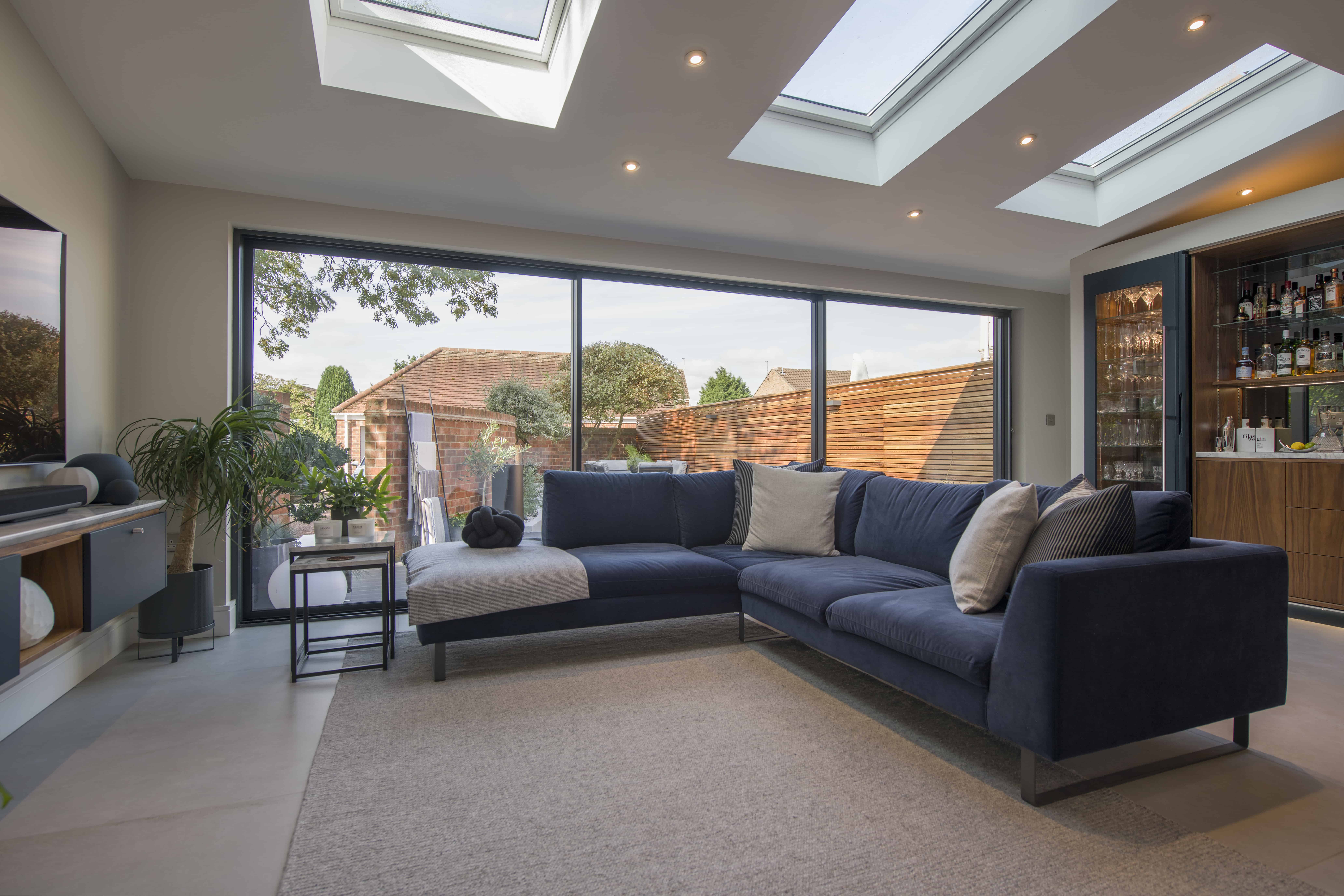 A modern living room with a navy sectional sofa, large skylights, and bifold doors opening to a garden. There is a built-in bar, a potted plant, and a cat resting on the chaise lounge.
