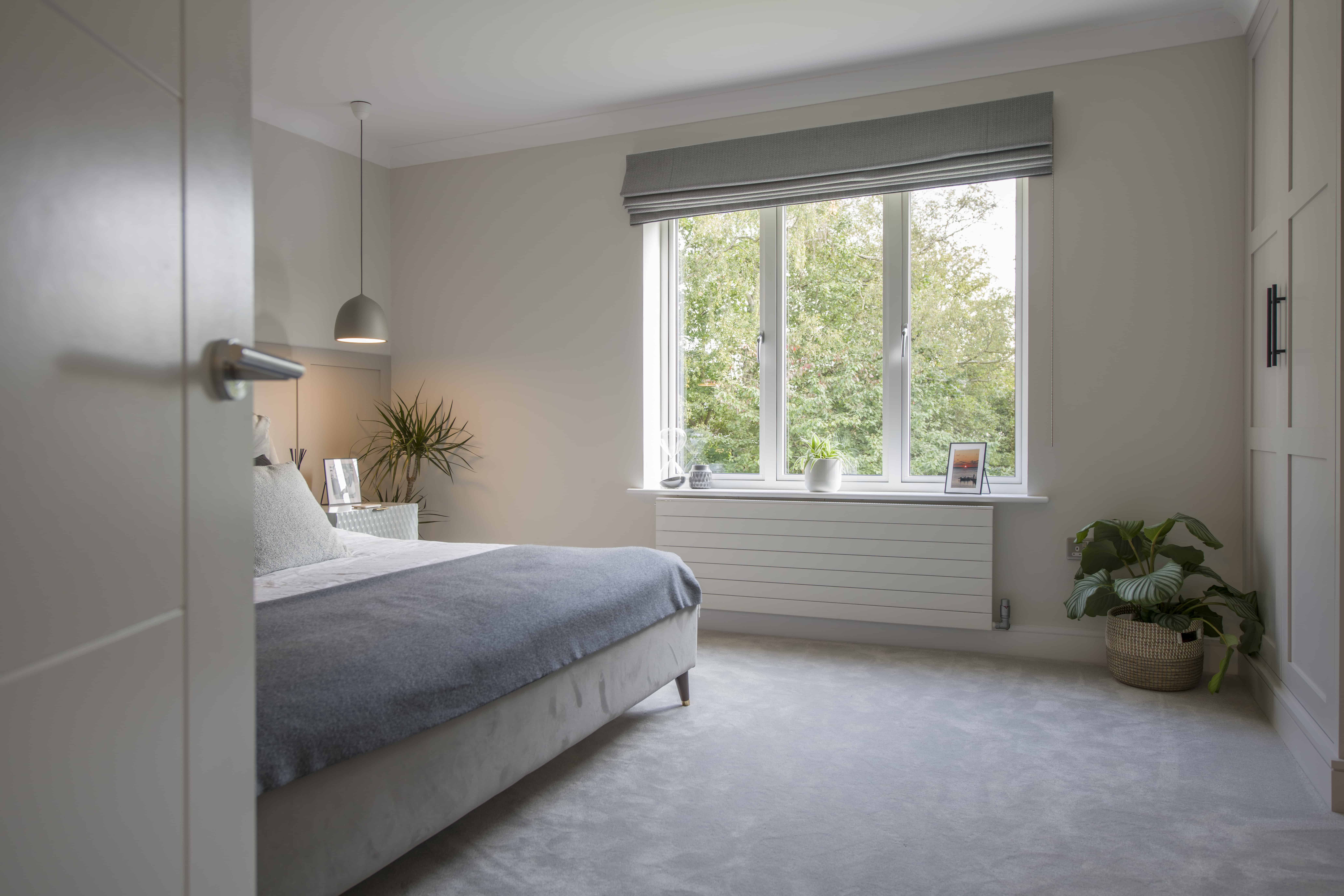 A bright, modern bedroom with a large window, sliding doors, a gray bed with white bedding, a potted plant by the window, a pendant light, and soft gray carpet. Trees are visible outside the window.