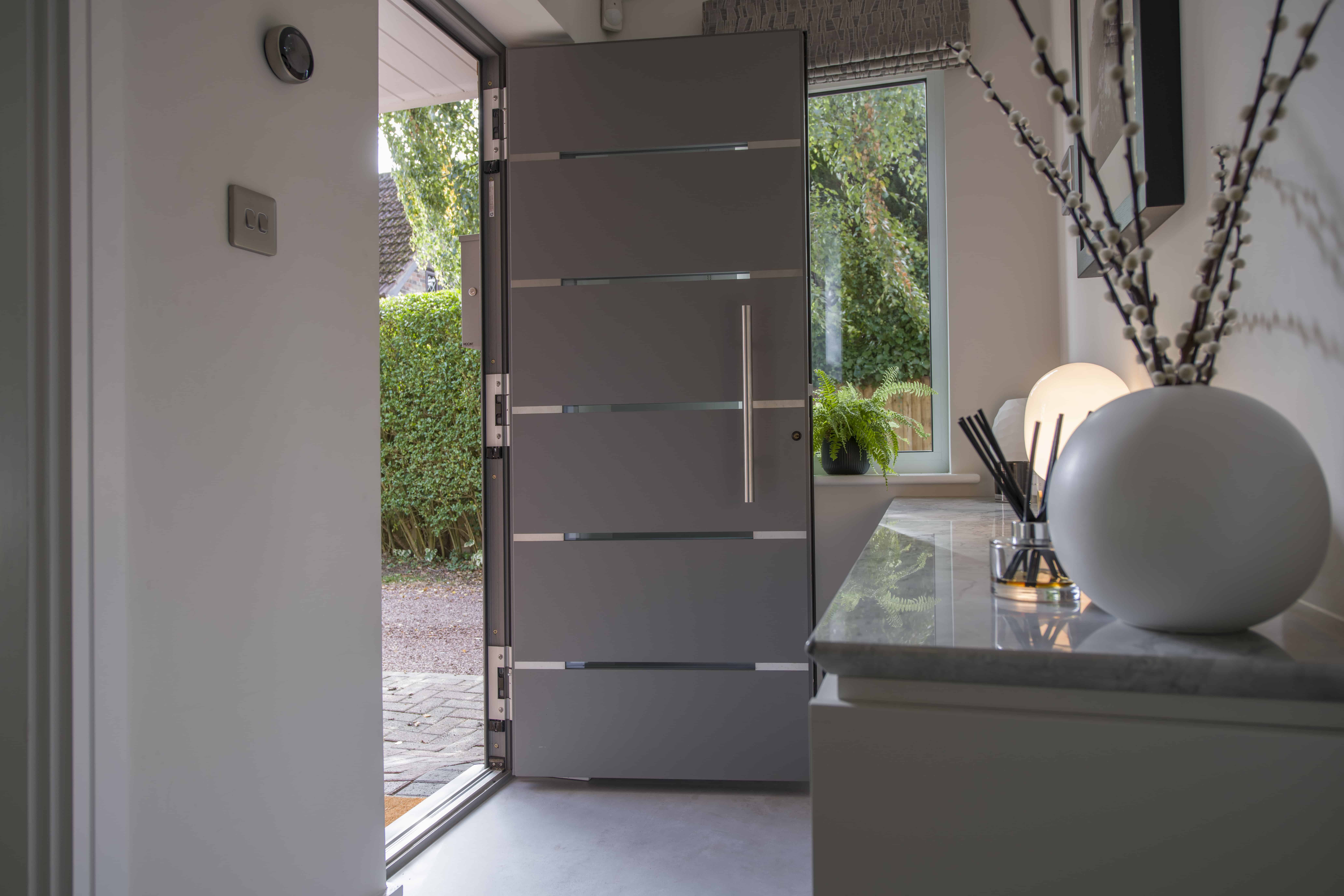 A modern entryway with a sleek gray front door partially open, letting in natural light. The interior features a marble-topped console with decorative items and a vase of branches. Bespoke glass solutions frame the view of lush greenery outside.