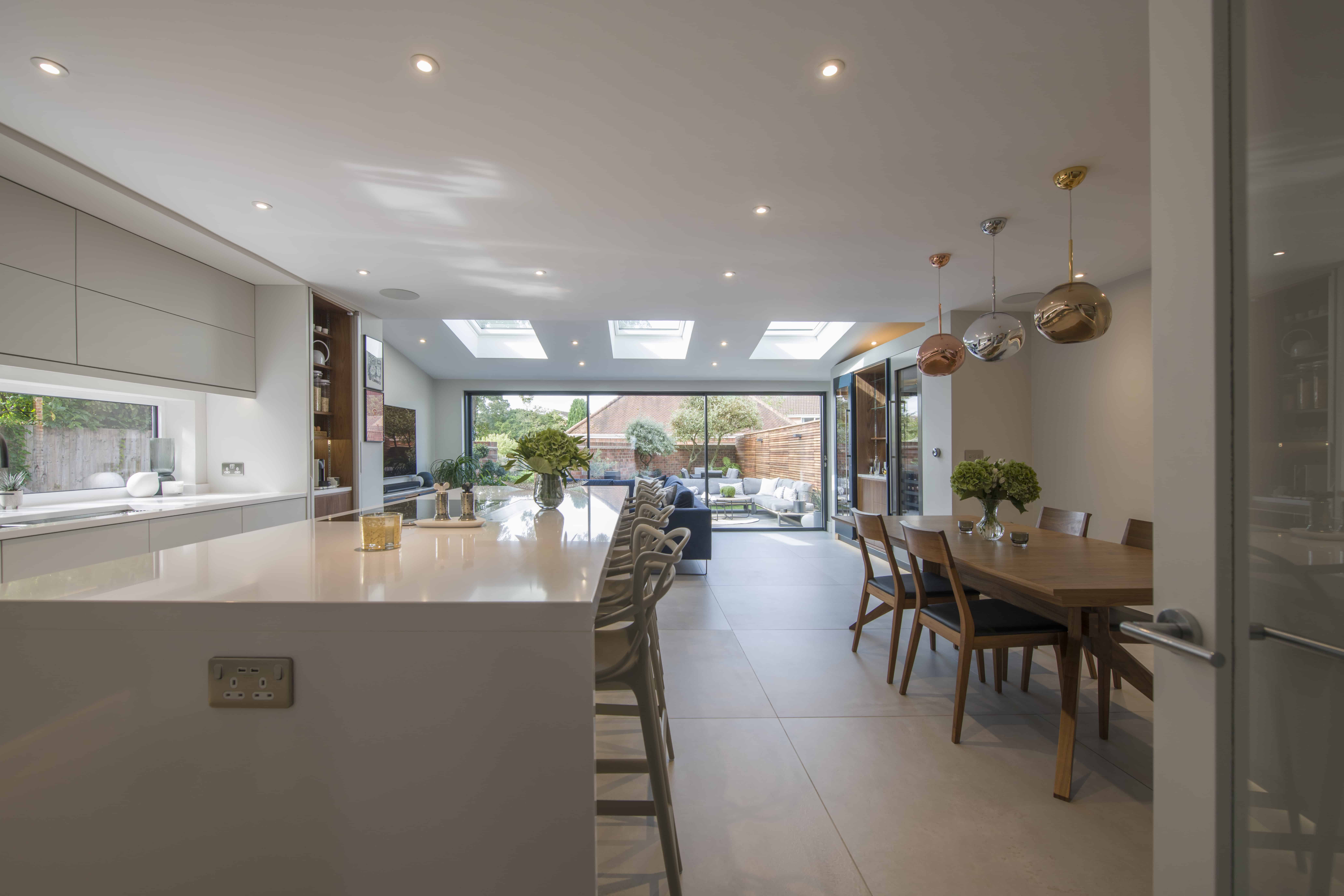 Modern, spacious kitchen and dining area with large island, bar stools, dining table, pendant lights, and bespoke glass solutions including sliding doors opening to a bright backyard with patio and garden.