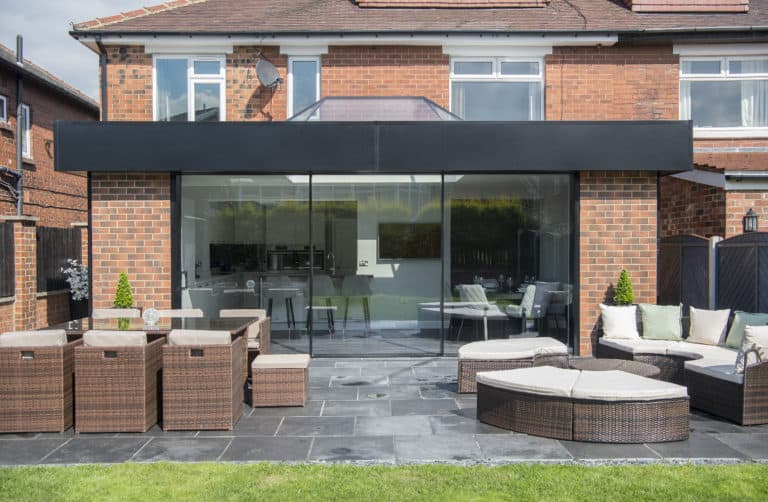 Modern brick house with large black-framed sliding doors opening to a patio with wicker furniture and a lawn. The patio features tables, chairs, white cushions for outdoor seating, and partial glass roofing for added style.