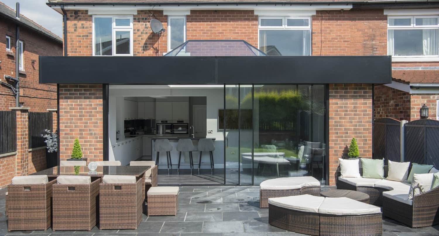 Modern patio with wicker furniture and cushioned seating set on gray stone tiles, in front of a brick house with large sliding doors opening to a contemporary open kitchen and bar area inside.