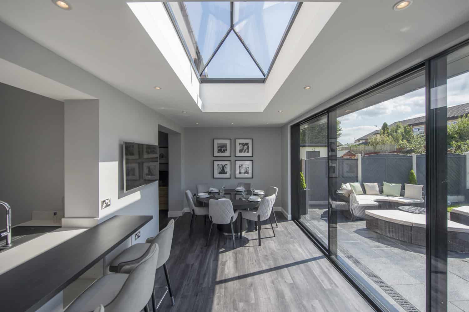 Modern, bright dining and kitchen area with a skylight, grey chairs, a round dining table, and large sliding bifold doors opening to a patio with round outdoor seating and a garden view.